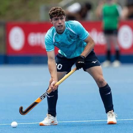 Field hockey player in blue jersey crouches on blue turf, gripping a stick near the ball, with a red banner in the background.