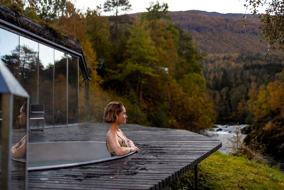 Woman relaxes in an outdoor hot tub beside a glass cabin, overlooking autumn mountains and a river in a peaceful forest setting