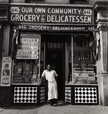 Man in apron stands outside "Our Own Community Grocery & Delicatessen." Black-and-white checkered tiles, vintage storefront of a black business in Harlem, New York.