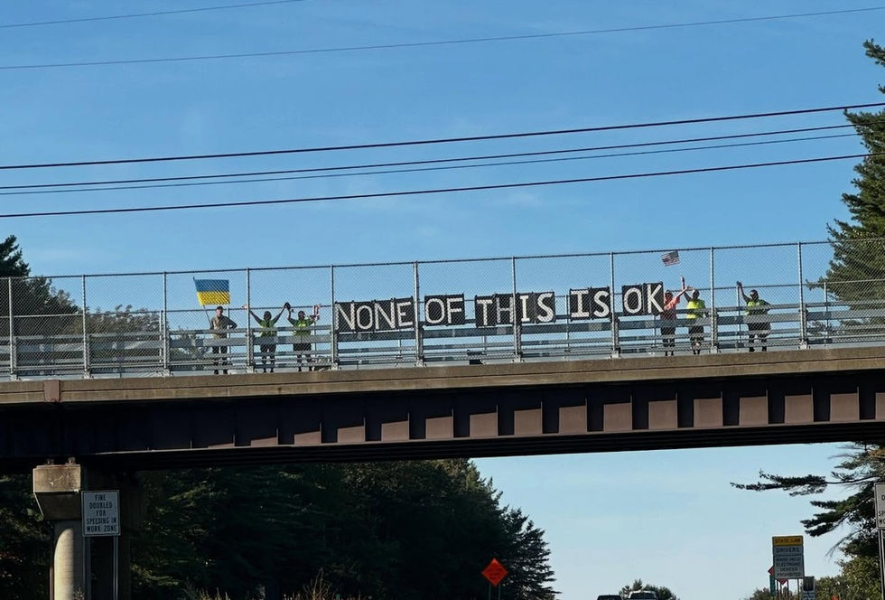 Peaceful New Hampshire Bridge Brigade volunteers holding a large banner reading “None of this is OK” on a highway overpass during a pro-democracy action