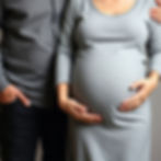 A clothed pregnant woman next to her husband. He has his arms around her. Cropped to just