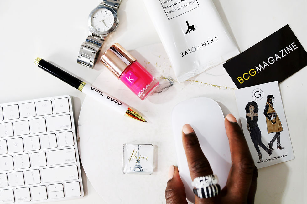 De cima vemos de forma desordenada e aleatória um teclado, uma caneta, um relógio, um hidratante, um cartão de visita, um esmalte e uma mãe negra segurando um mouse