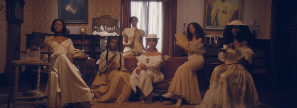 Beyoncé está sentada numa sala, rodeada de várias outras mulheres negras, todas com roupas vitorianas, penteados tradicionais da comunidade afro americana, e ar de quem manda