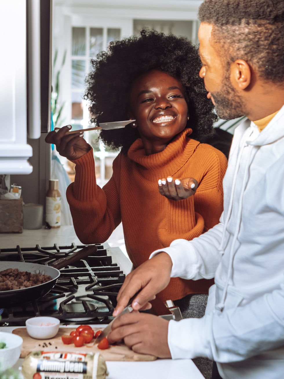 Um homem e uma mulher, ambos negros, cozinhando juntos. Ela está sorrindo e oferecendo um pouco de comida para ele experimentar