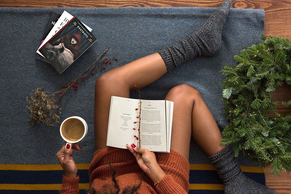 Imagem vista de cima, uma pessoa negra está sentada em cima de uma manta, segurando uma caneca de café em uma mão e um livro aberto na outra. No chão mais livros e uma guirlanda apenas de folhas verdes