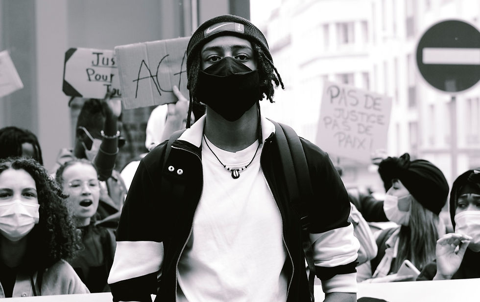 Um jovem negro está no centro da imagem, no meio de um protesto. Está usando máscara e olhando seriamente para a câmera