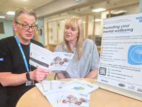 A Wellbeing for Life Practitioner is talking to a client and looking through a service leaflet. There are workbooks and a roller banner on the table where they are sast