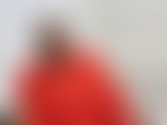 A lady is sat looking directly at the camera. She is wearing bright clothes, a head scarf and glasses.