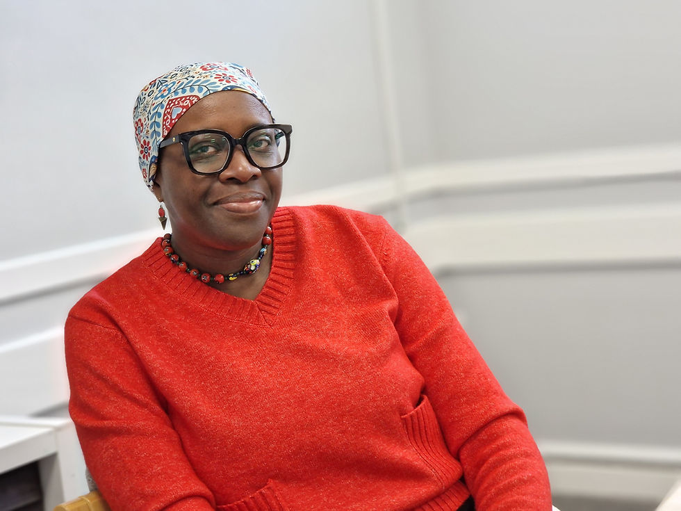 A lady is sat looking directly at the camera. She is wearing bright clothes, a head scarf and glasses.