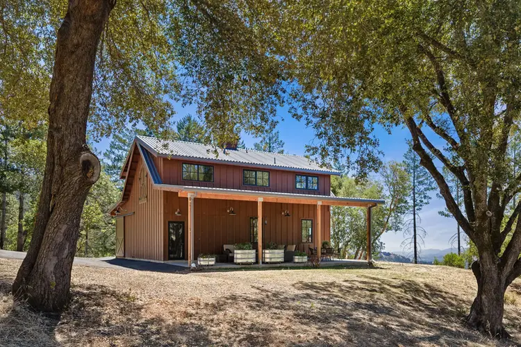 Rustic barn-style home with metal roof and covered front porch, set among trees on a gravel drive.