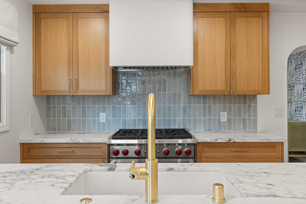 Cooking wall with rift white oak cabinets, white range hood, blue tile backsplash, and brass faucet over a large marble-look island.