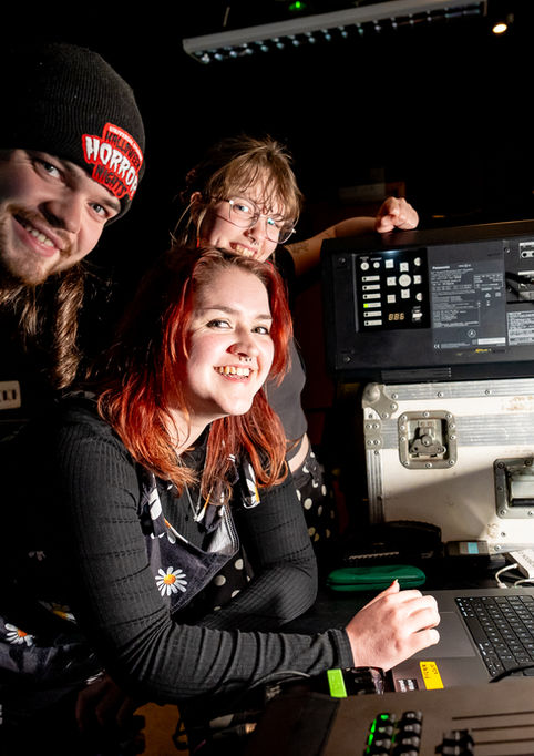 A photo of the video team standing over a laptop and projector. 3 people, all looking to the camera and smiling