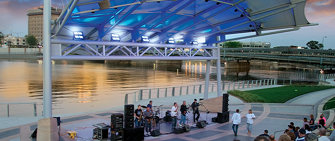 A band performs for a crowd next to the Cedar River under an artistic-looking overhang at the downtown Waterloo amphitheater.