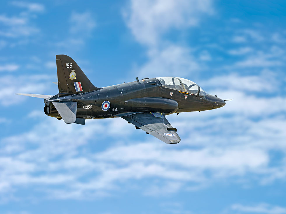 A black military jet soars against a blue sky with white clouds. Markings on tail and fuselage include "156" and "XX156."