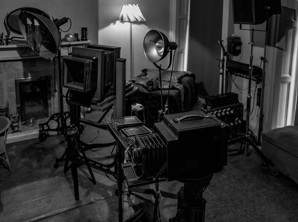 Black-and-white photo studio with vintage large-format cameras, lights, and stands in a dim room, evoking a moody workspace