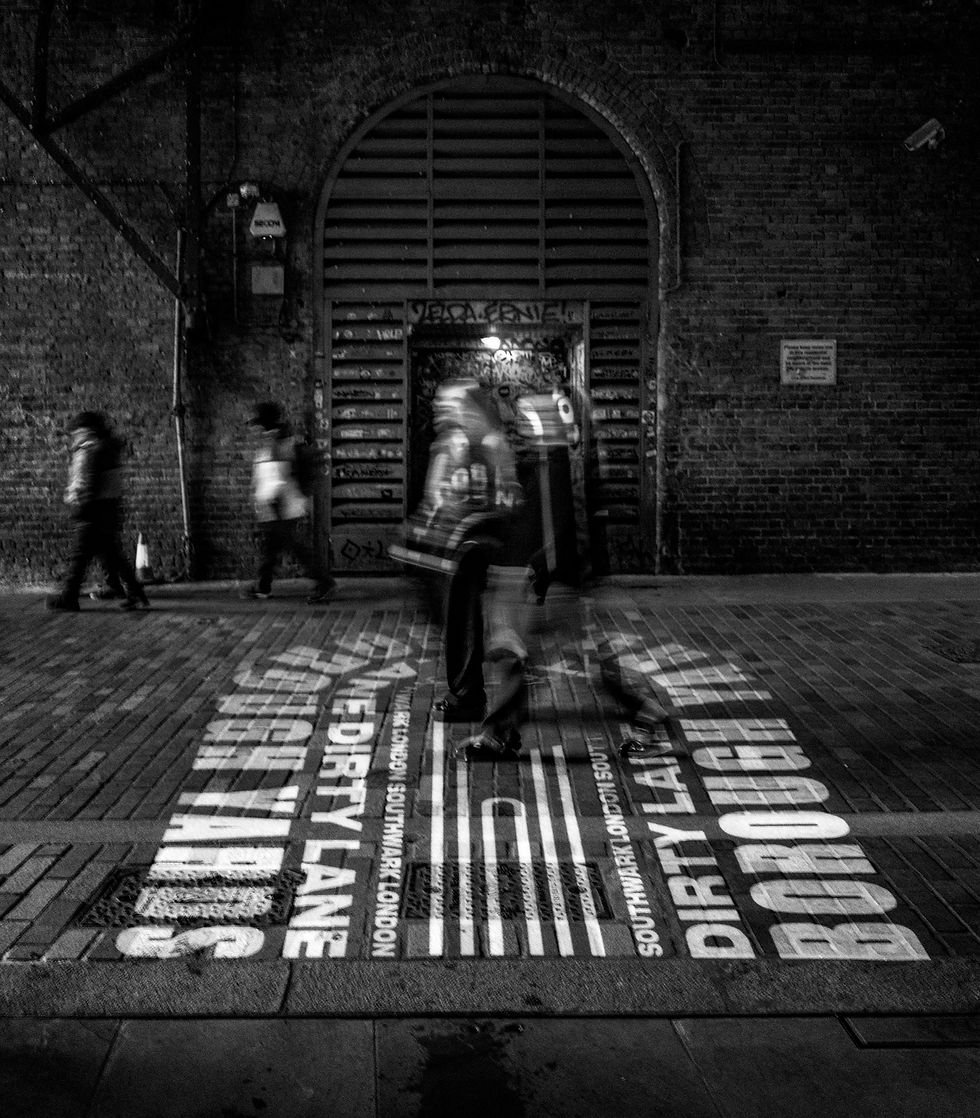People walking on a brick street under an arched doorway with graffiti. "Dirty Lane Borough Yards" text on ground. Black and white mood.
