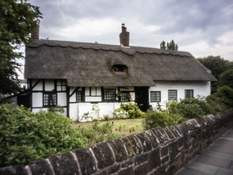 Thatched cottage with white walls and black timber trim behind a stone wall, under a cloudy sky with lush greenery