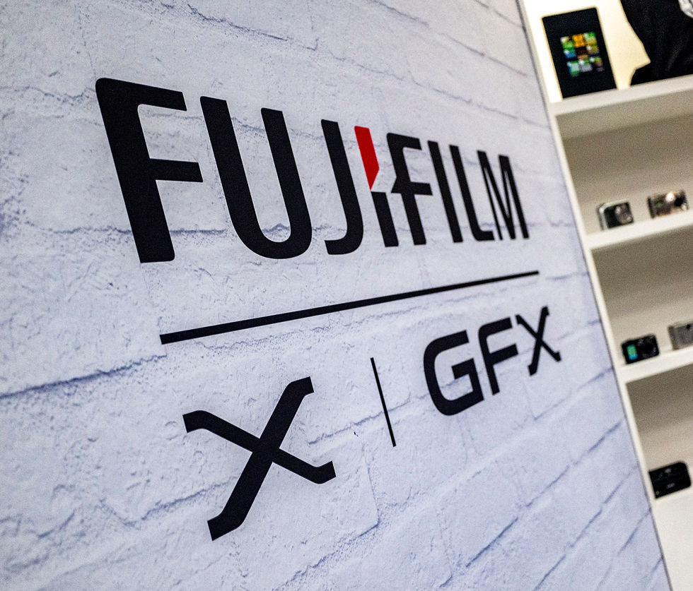 FUJIFILM X GFX logo on a white brick wall. Shelves with cameras in the background. Bold black and red typography, tech-themed setting.