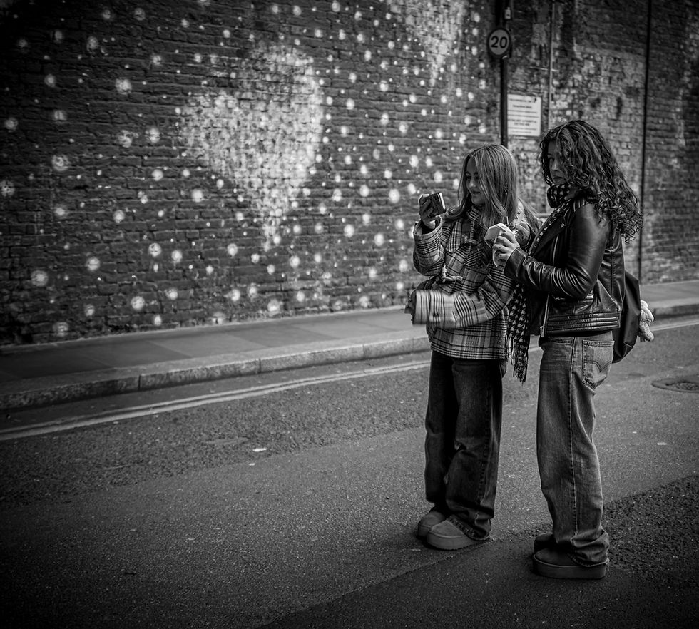 Two people on a street take a photo in front of a heart mural on a brick wall. They appear casual and relaxed, holding drinks.