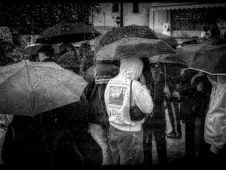 Crowd in rain with umbrellas, seen through wet glass. Monochrome mood. Text on a hoodie reads "The Field with Open Eyes" and "Vincent Van Gogh".