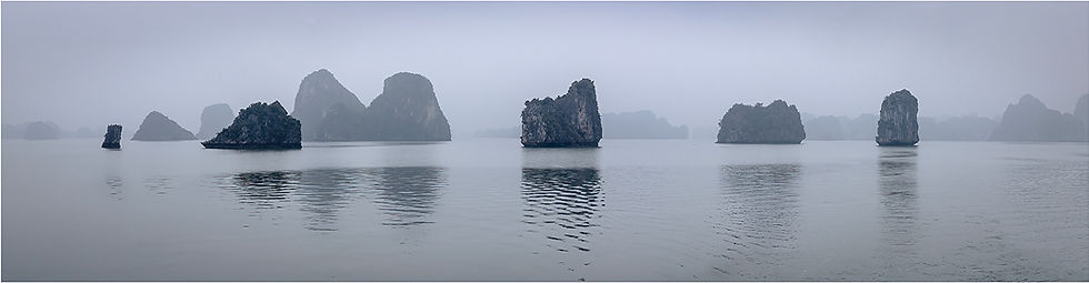 Mist-covered limestone islands rise from calm waters under a gray sky, creating a serene and tranquil seascape.