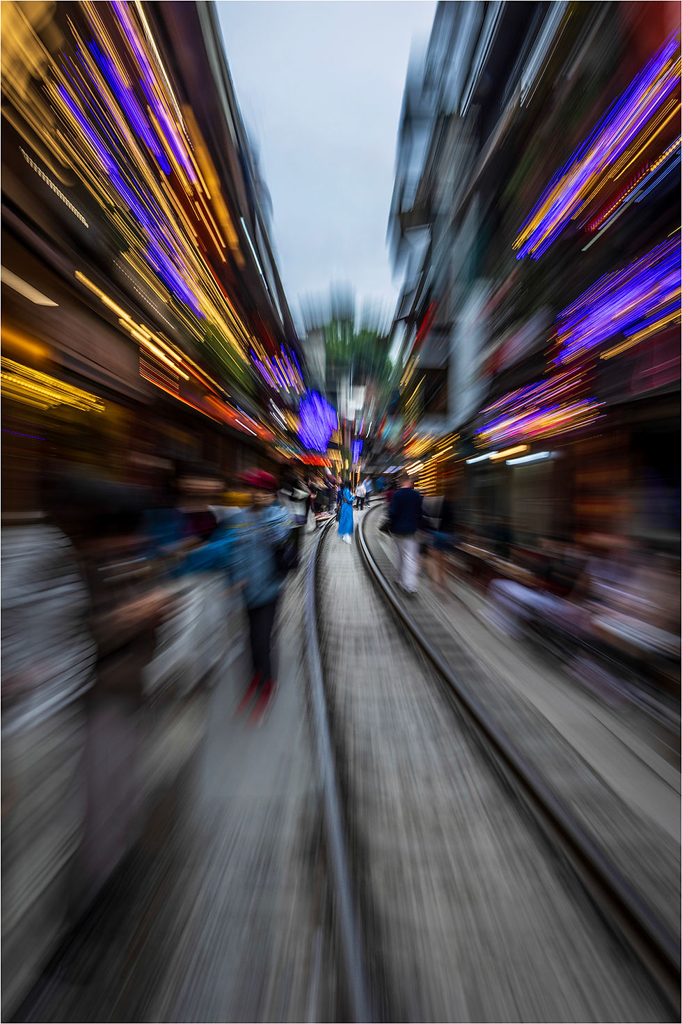 Blurred street view with vibrant blue and yellow lights. People walk along a railway track, creating a dynamic motion effect.