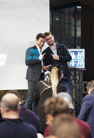 Two men in suits standing on a stage, smiling and pointing toward a small display screen while an audience watches from below.