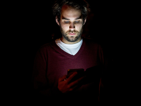 Man illuminated by a phone screen in a dark room, representing isolation and digital addiction.