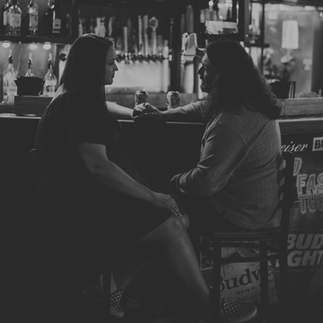 A couple embraces amidst the dimly lit ambiance of Cool Beans Bar & Grill, their smiles radiant in the cozy atmosphere. In another photo, they share a tender moment, bathed in the warm glow of the bar's neon lights. Each image captures the raw authenticity and deep connection between the couple as they celebrate their love in this unique setting.