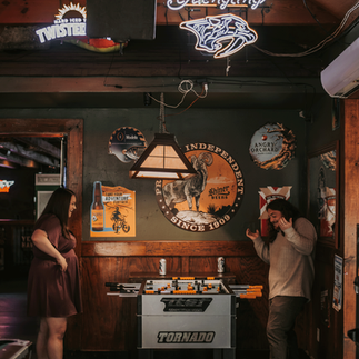 A couple embraces amidst the dimly lit ambiance of Cool Beans Bar & Grill, their smiles radiant in the cozy atmosphere. In another photo, they share a tender moment, bathed in the warm glow of the bar's neon lights. Each image captures the raw authenticity and deep connection between the couple as they celebrate their love in this unique setting.