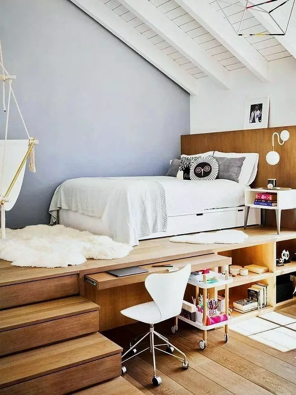 Stylish small bedroom with stairs leading up to a bed, next to it is a office chair that is white sitting under a desk connected to the bed.