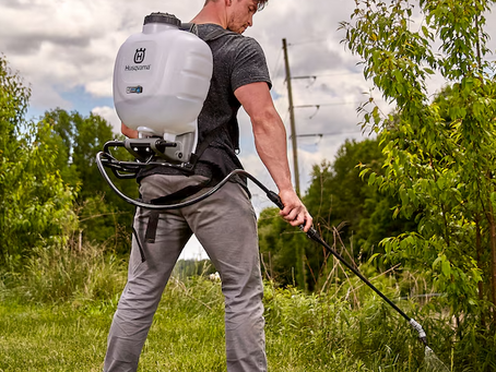 Man spraying plants with a backpack sprayer outdoors. Cloudy sky, green trees, and grass in the background. Husqvarna logo visible.
