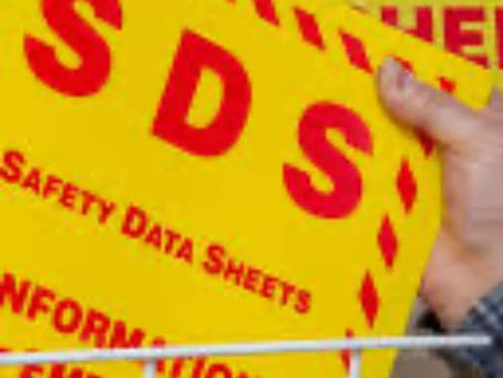 Hand placing a yellow SDS safety sign with red text on a rack. Background shows hazard symbols and text. Industrial setting.