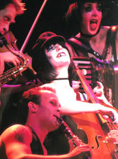Cello player smiles in a theatre during the show Cabaret, and is surrounded by otherr musicians including a clarinettist and accordion player. Margit van der Zwan plays the cello