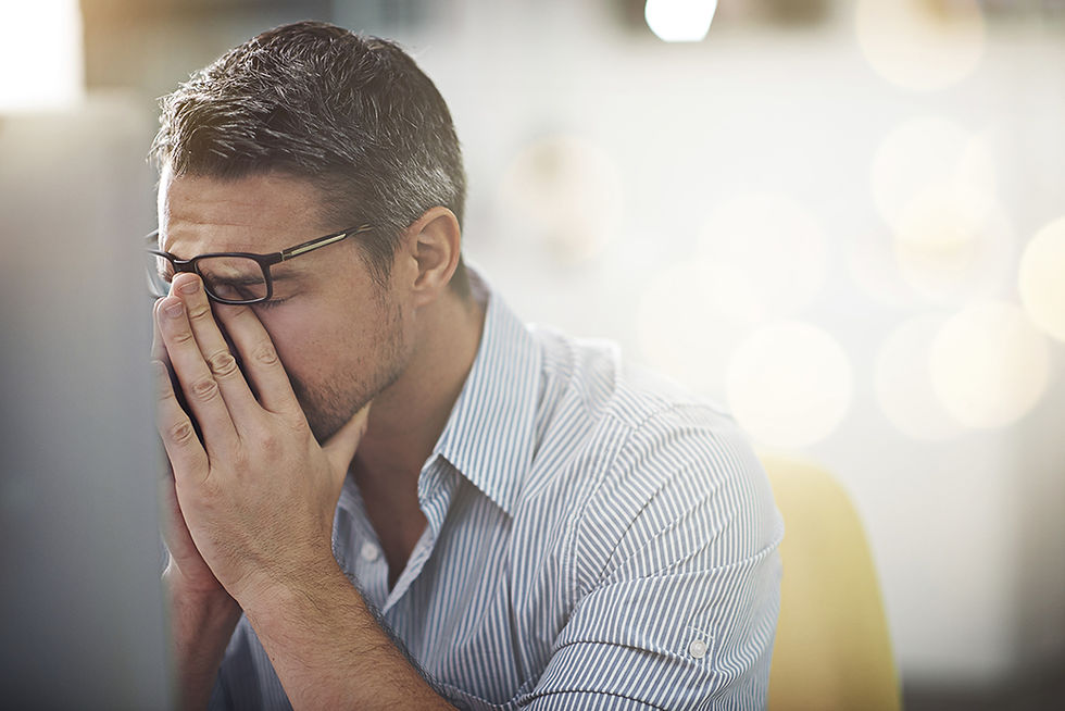 Man with glasses and striped shirt sits with hands on face, looking stressed in a blurred office setting with soft lighting.