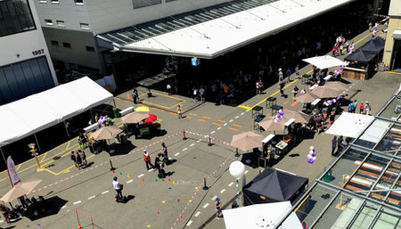 Menschen besuchen einen Aussenmarkt mit Zelten und Ständen unter Gebäuden.