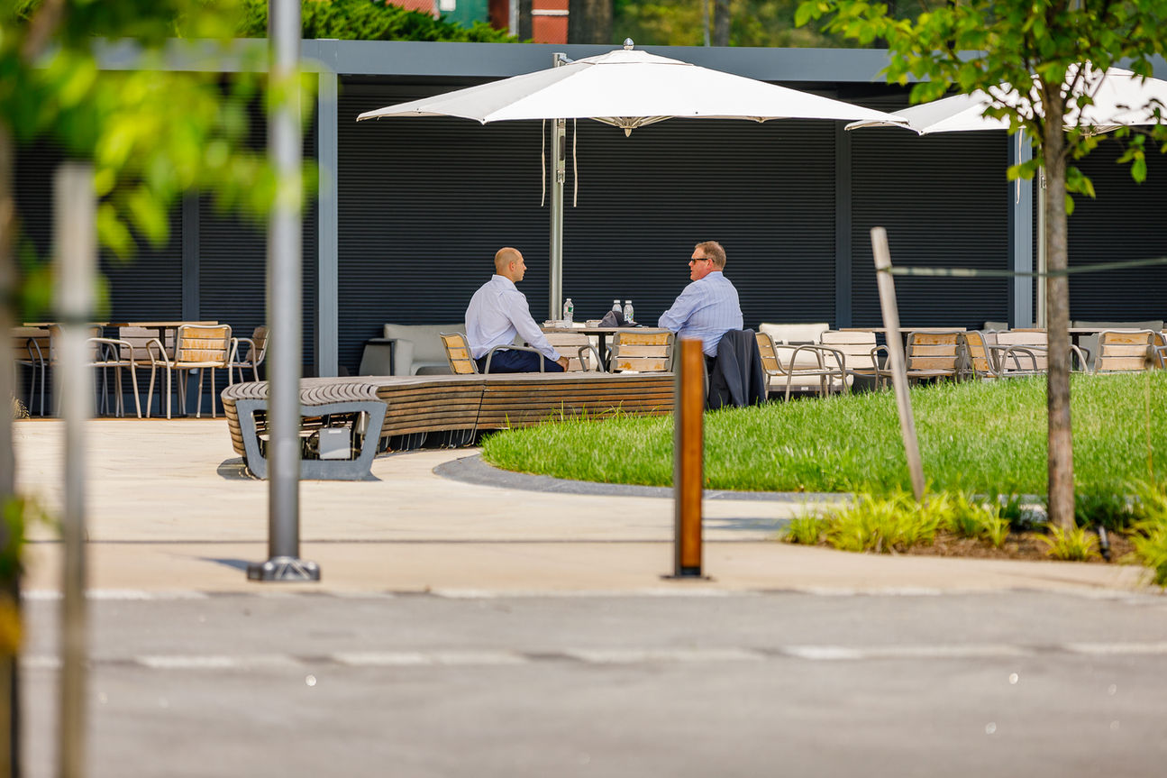 Two business professionals having a meeting at an outdoor table under a white umbrella, adjacent to a curved wooden bench and green lawn.