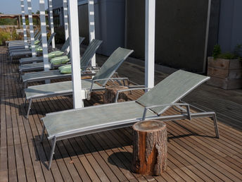 A row of modern metal lounge chairs with green cushions on a wooden deck beneath a white pergola structure, accented with rustic tree stump side tables.