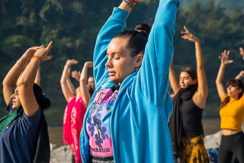 Group of women practicing outdoor movement or yoga on a sandy riverbank, led by a woman in a bright blue hoodie and black shorts with her arms raised overhead; participants mirror the pose behind her, surrounded by lush green forested hills and mountains, capturing a wellness retreat, mindful movement, and nature-based fitness experience.