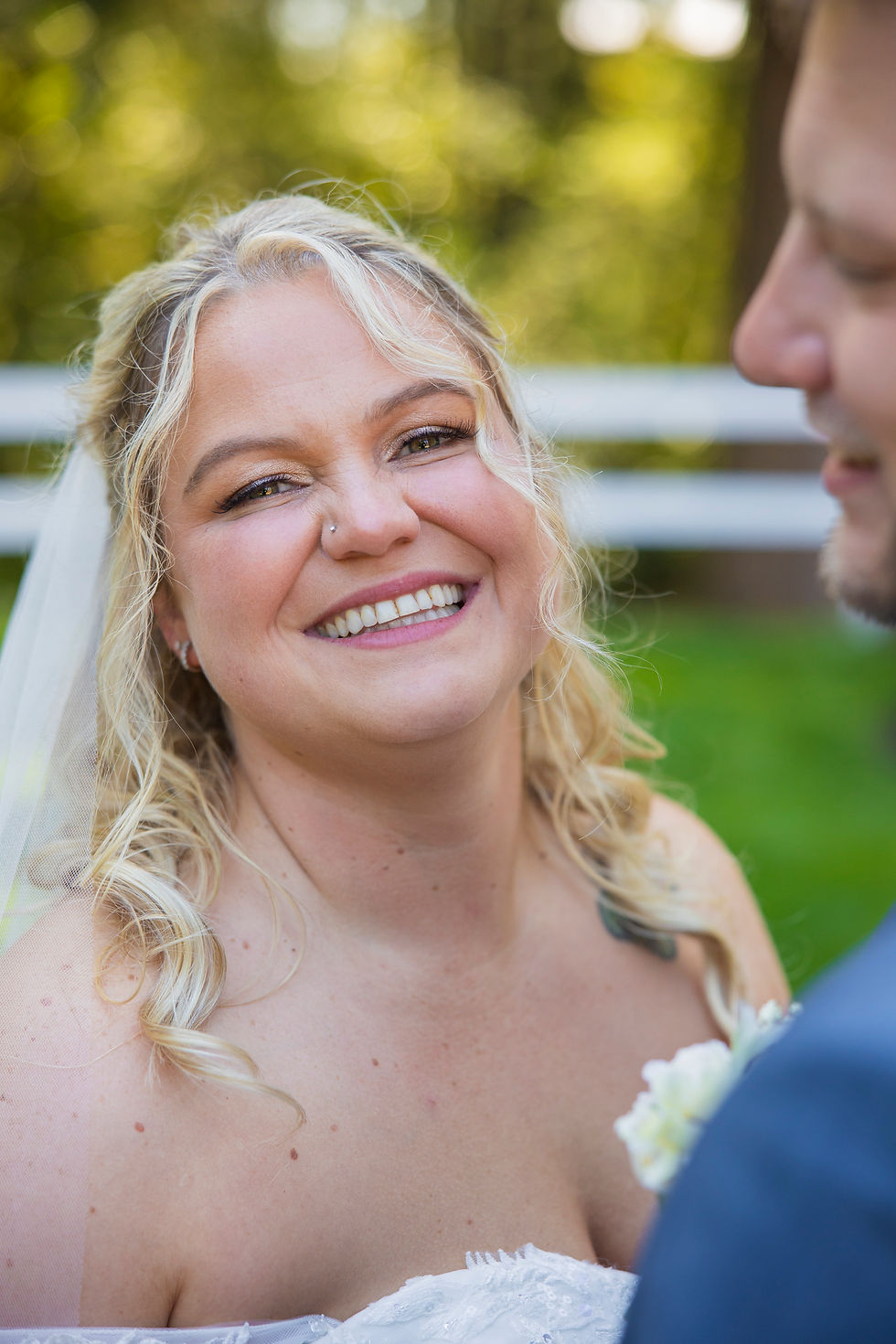 close up portrait of Bride in white dress smiling joyfully at groom outdoors. Green background, sunny setting at golden hour, showing happiness and love.