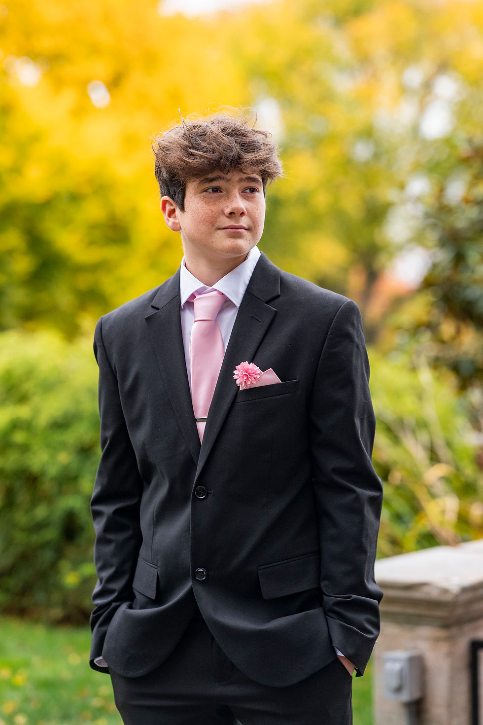 Young man in a black suit with a pink tie and flower stands in a park, hands in pockets. It's fall leaves in background in Tacoma WA for Homecoming dance