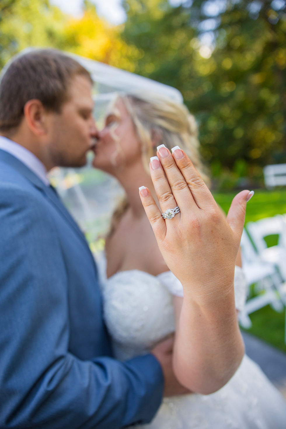 Bride and groom kiss outdoors under veil portrait by Sage and Soul Photography; bride's hand with diamond ring in focus. Sunlit garden setting and joyful atmosphere.