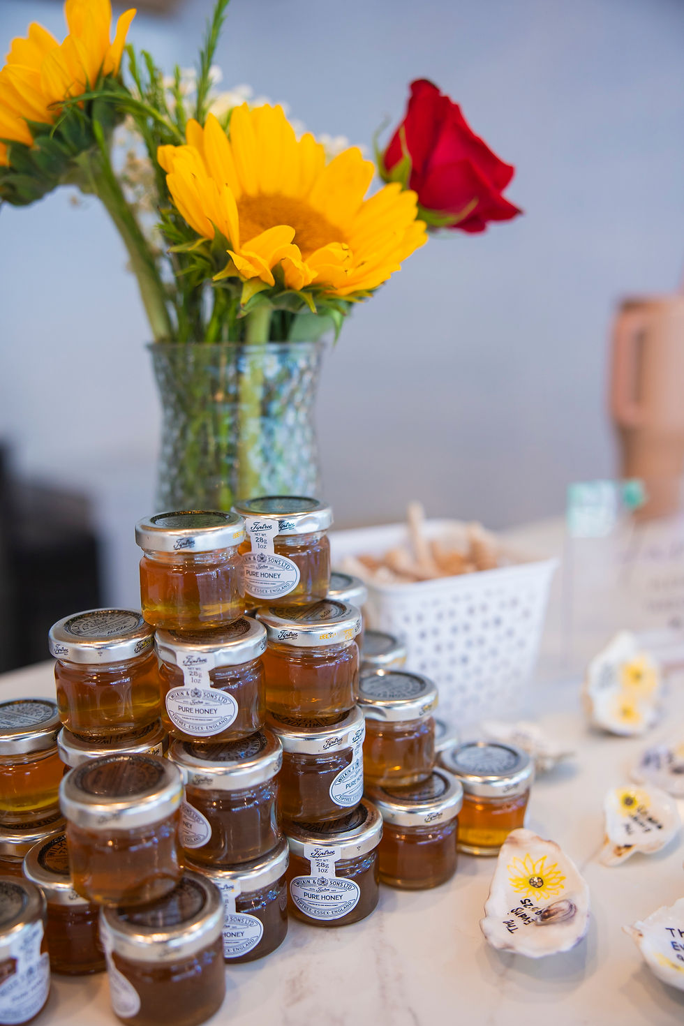 Small jars of honey stacked on a table with vibrant sunflowers and a red rose in a vase nearby, creating a cheerful setting.