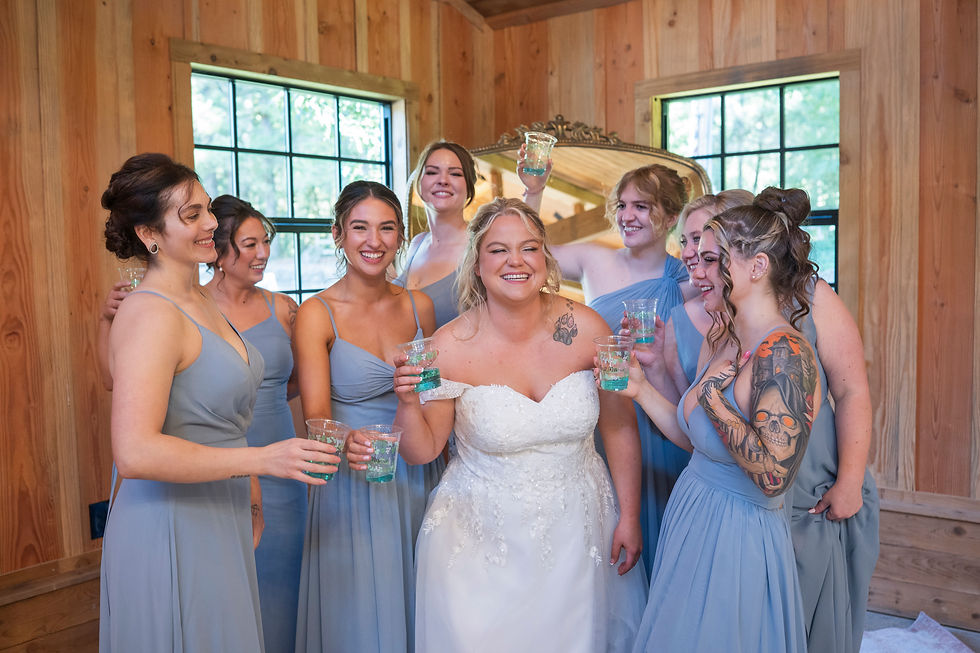 Bride in a white dress with six bridesmaids in mismatched blue, all smiling and toasting in a wooden room. Bright window light enhances the joyful mood in barn wedding
