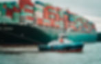 Tugboat pulling large cargo ship stacked with multicolored containers labeled "EVERGREEN." Overcast sky and calm water in the background.