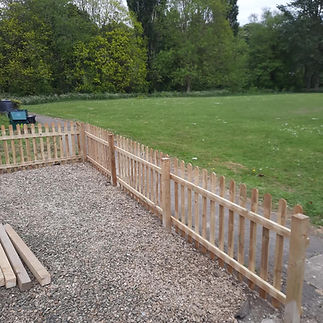 A new wooden picket fence separates a gravel area from a green grassy field