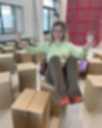 Woman smiling, sitting playfully on cardboard boxes, holding a wine glass. She's in a bright room with modern decor, wearing a green top.