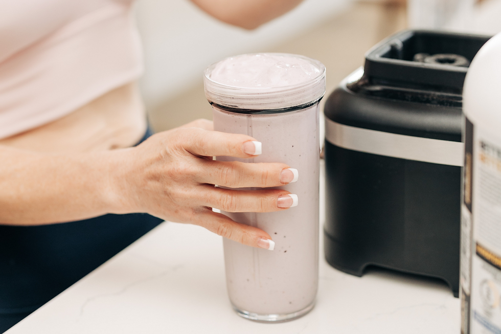 Hand holding a glass of purple smoothie near a black blender on a white counter. French manicure nails visible. Bright setting.
