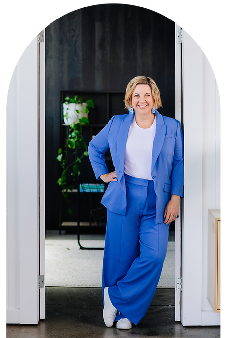 Tracey in a blue suit and white top, with dangly blue earrings. She is leaning against a wall, smiling, hand on hip.