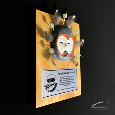 This is a depiction of a Birch award plaque finished in natural tones. It has a metal silver award plate with black lettering. Affixed above the award plate is a Yup'ik stylized Salmon maskette in grey, white, and orange colors. Porcupine quills with sea glass beads, and feathers, surround the artwork.